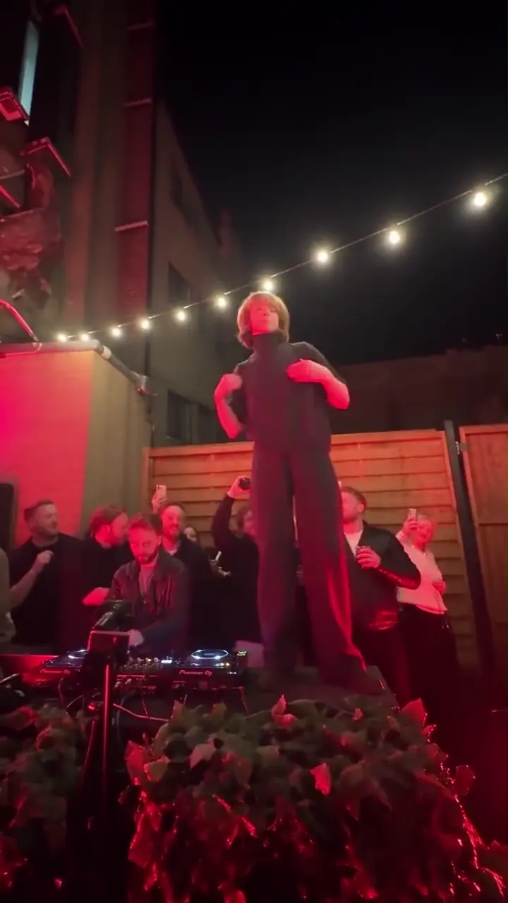A dancer with short blonde hair in a dark jumpsuit crosses both arms over the chest and flicks them out on the planter, red light behind and the DJ decks in front.
