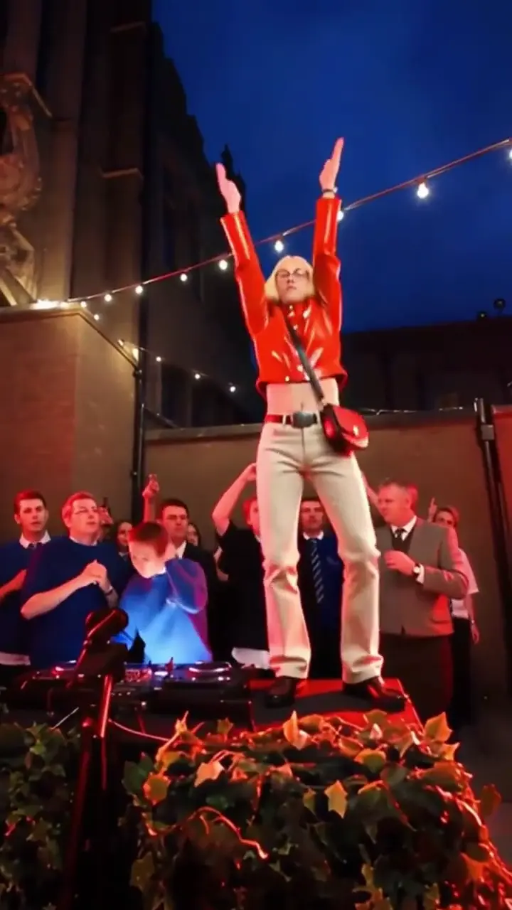 A dancer with platinum hair in a red leather jacket and white jeans rolls the wrists and points both hands out from the planter, the DJ booth lit up in front.