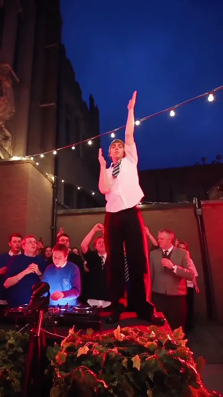 A man in a white shirt and a striped tie stands on the planter above the crowd and snaps his arms across his face and up over his head under the string lights.